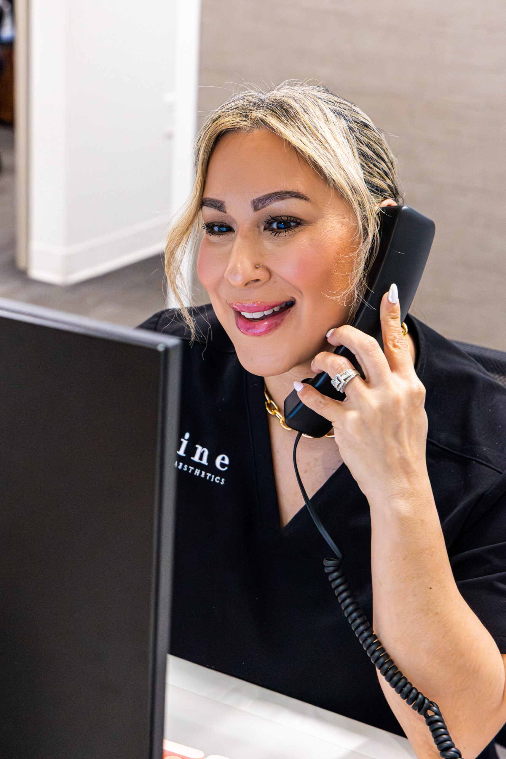A blonde front desk associate at Shine Medical Aesthetics in Alpharetta, GA, wears black scrubs while taking a phone call to schedule a cosmetic injectables consultation and appointment.